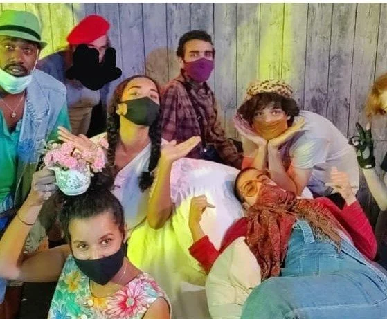 Menschen in einer Gruppe, alle tragen Masken und posieren für ein Foto, vor einer hölzernen Wand. Einige sind jung, einige älter, und sie scheinen eine fröhliche Stimmung zu haben.