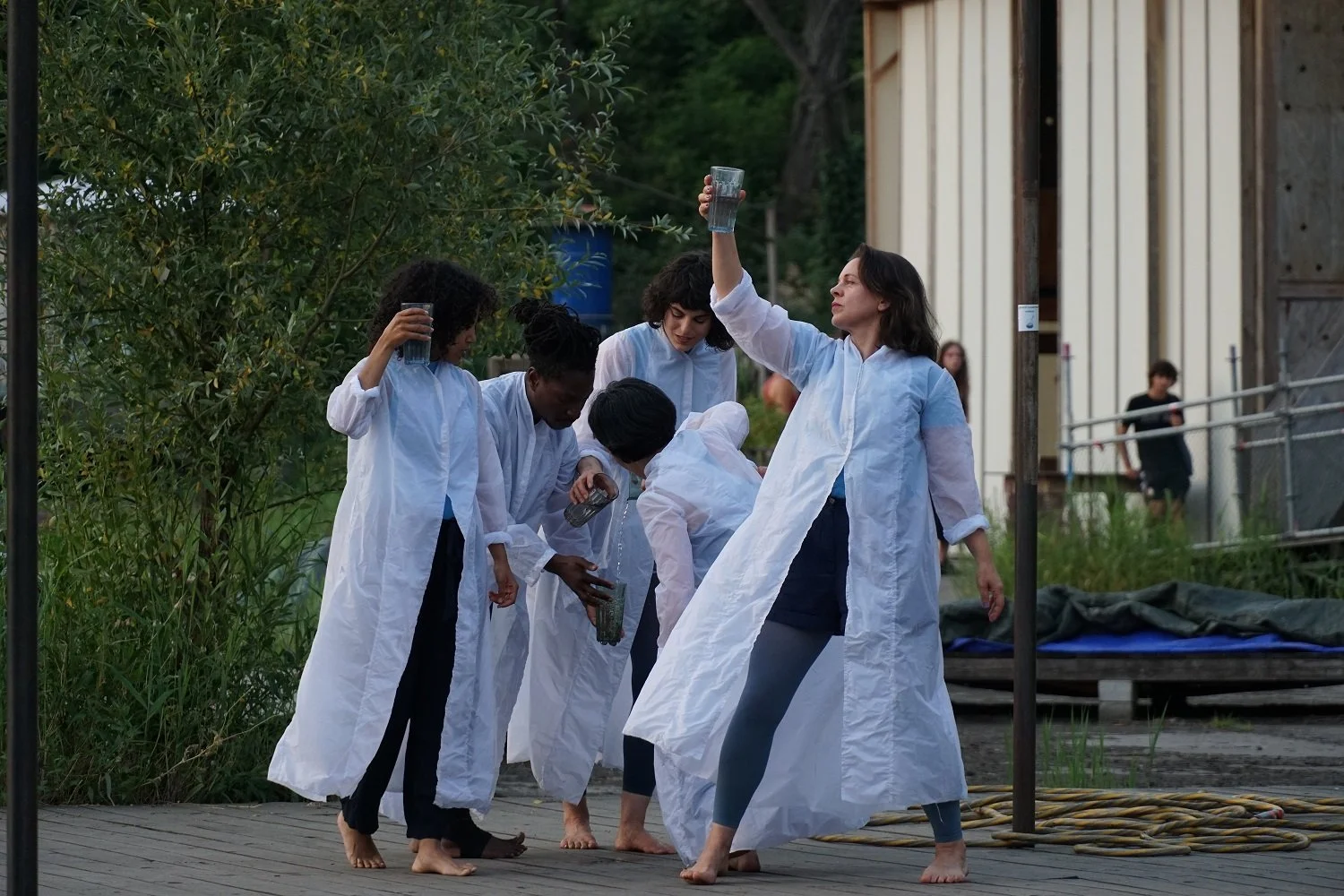 Eine Gruppe von Frauen in weißen Kitteln, die auf einer Holzplattform vor einem Pflanzenbereich stehen, scheinen an einem Outdoor-Aktionen teilzunehmen, mit einigen den Blick nach unten gerichtet, während eine Frau ihre Hand mit einem Glas hoch in die Luft hält.