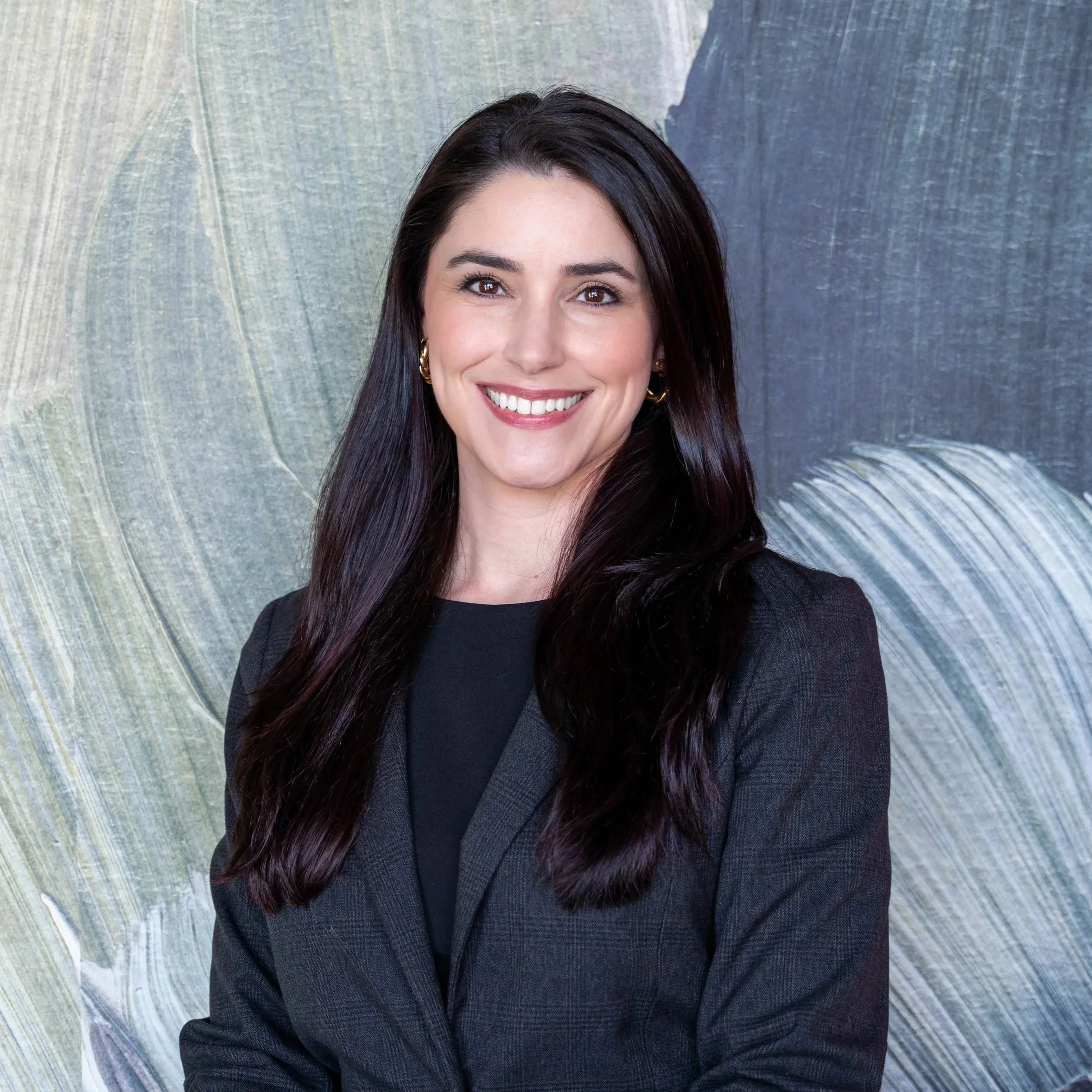 A woman with long dark hair and fair skin, wearing a black blazer, sitting on a pink couch with her hand resting under her chin, looking directly at the camera.