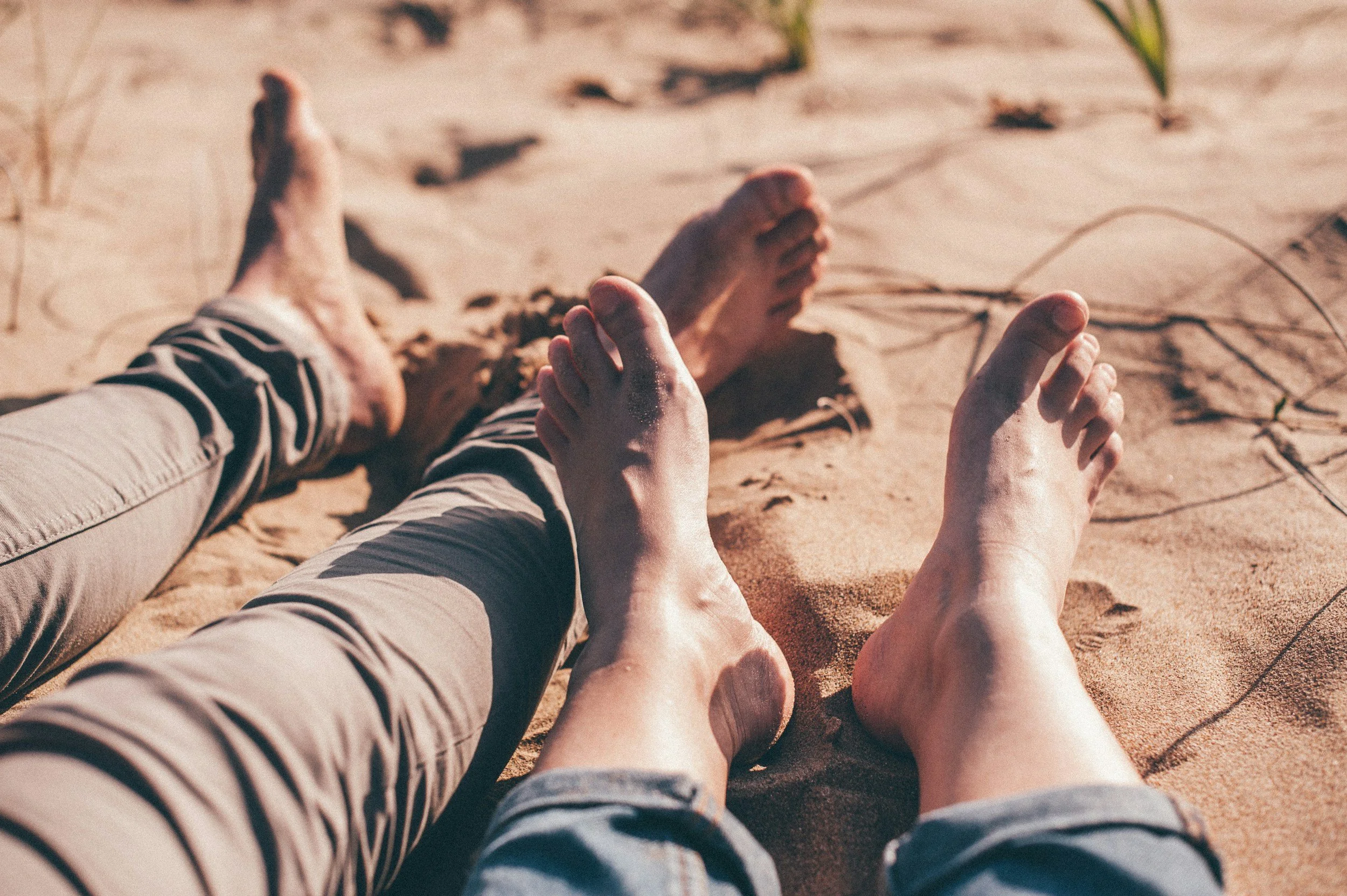 Two pairs of legs and feet lying on sandy ground at the beach, with some grass and twigs nearby.