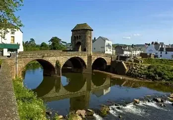 A historic stone bridge over a river with a small tower and houses in the background, reflecting in the water.