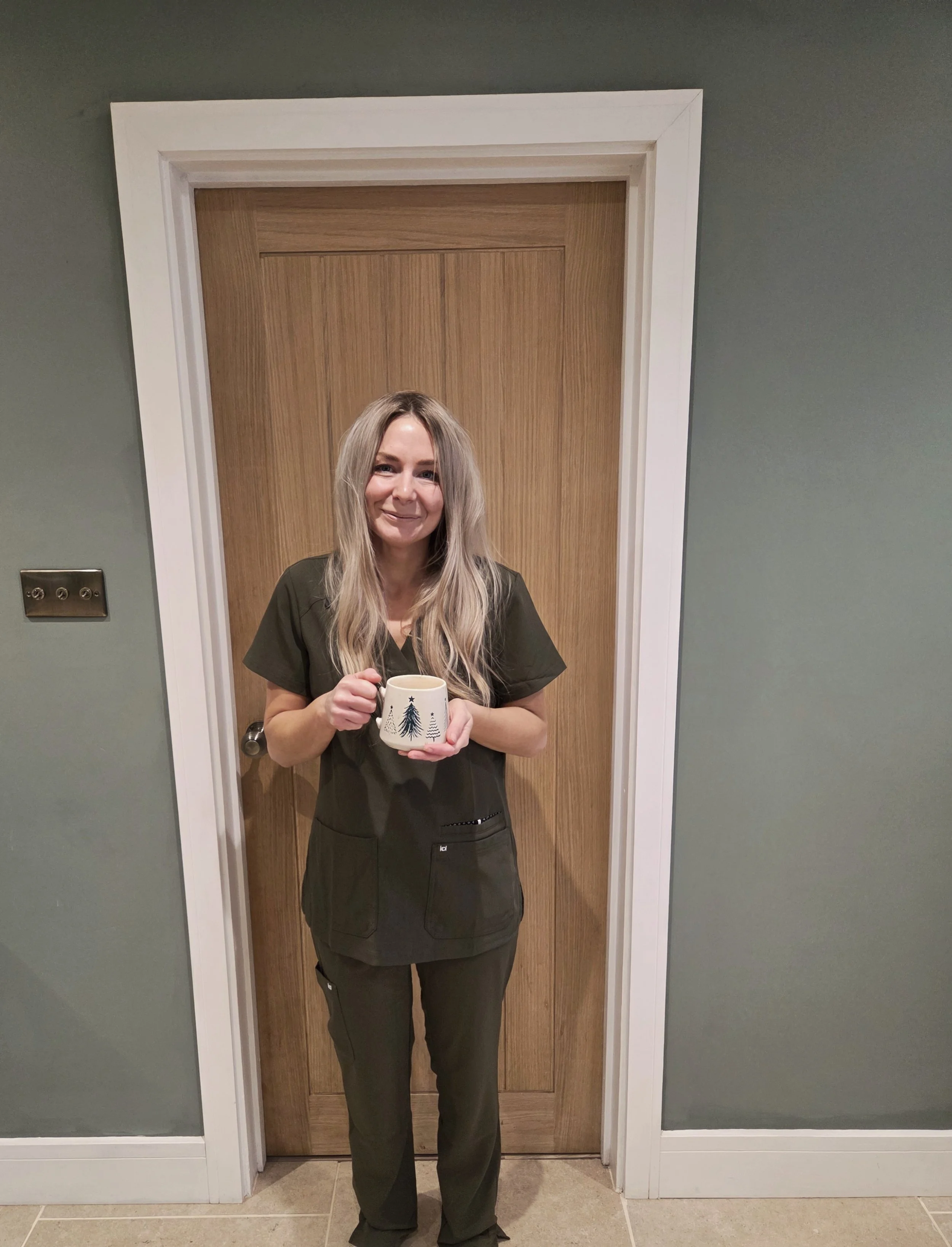 A woman with long blonde hair smiling and holding a mug with a Christmas tree design, standing in front of a wooden door in a green-walled room.
