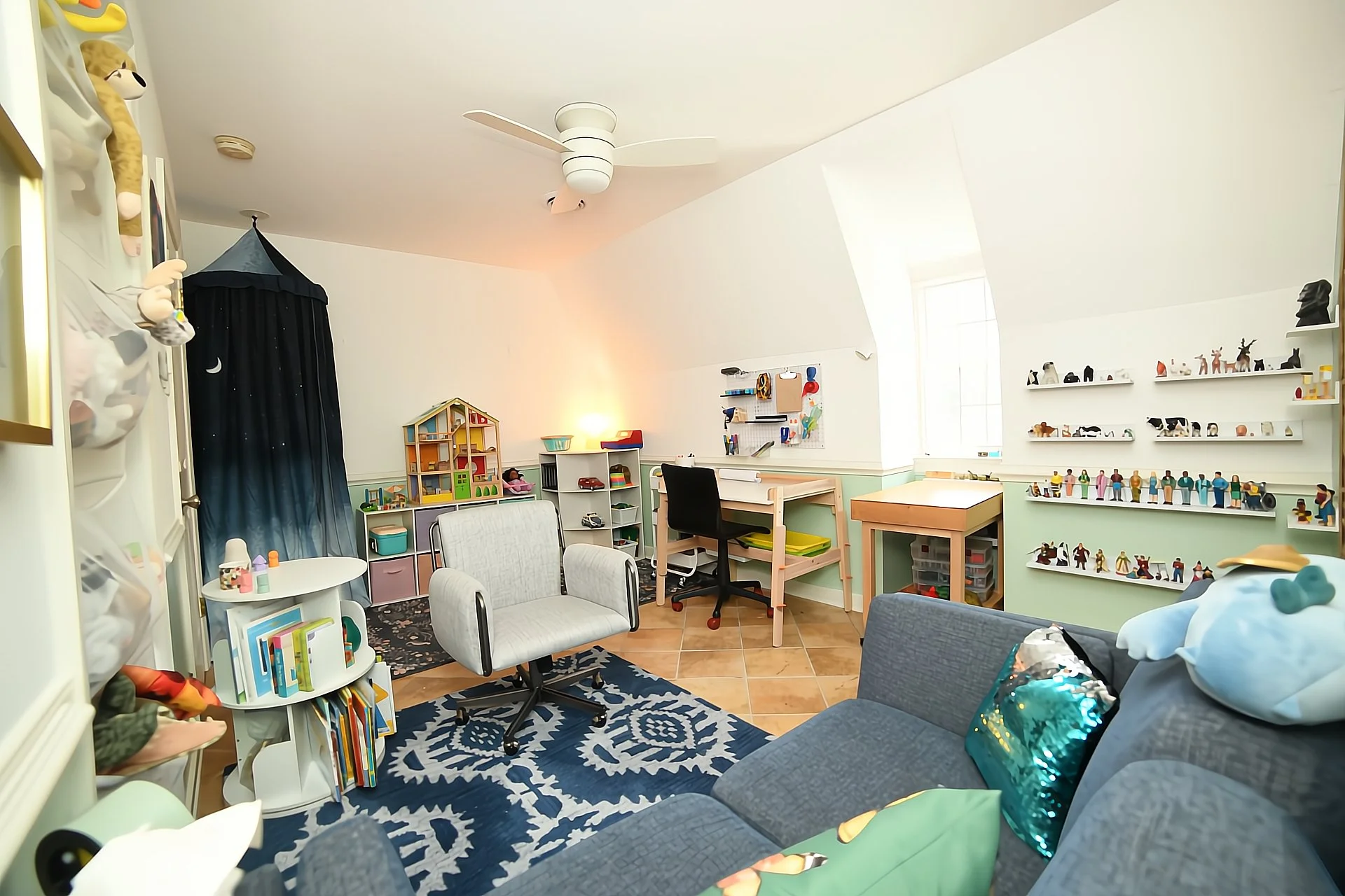 Therpeutic playroom with various toys, sand tray, dollhouse, and decor, including a blue patterned rug, white chairs, a small art table, and a blue sofa. Toys and decorations are scattered around.