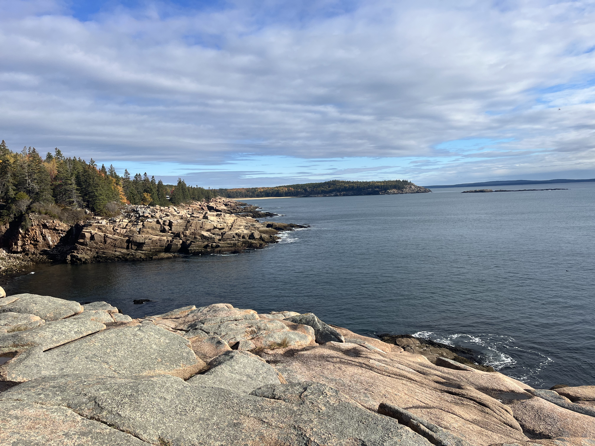 Rocky shoreline with trees on the left and calm water extending to distant islands under a partly cloudy sky.