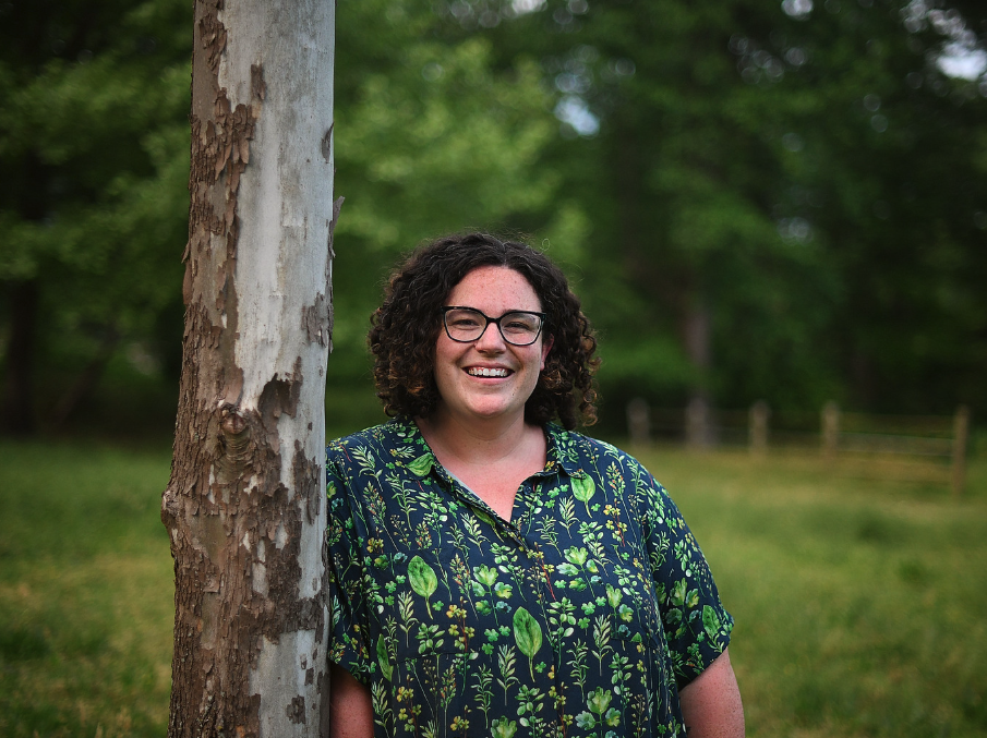 A woman with curly dark hair, glasses, and a floral shirt leaning against a tree outdoors, smiling at the camera with a blurred green landscape in the background.