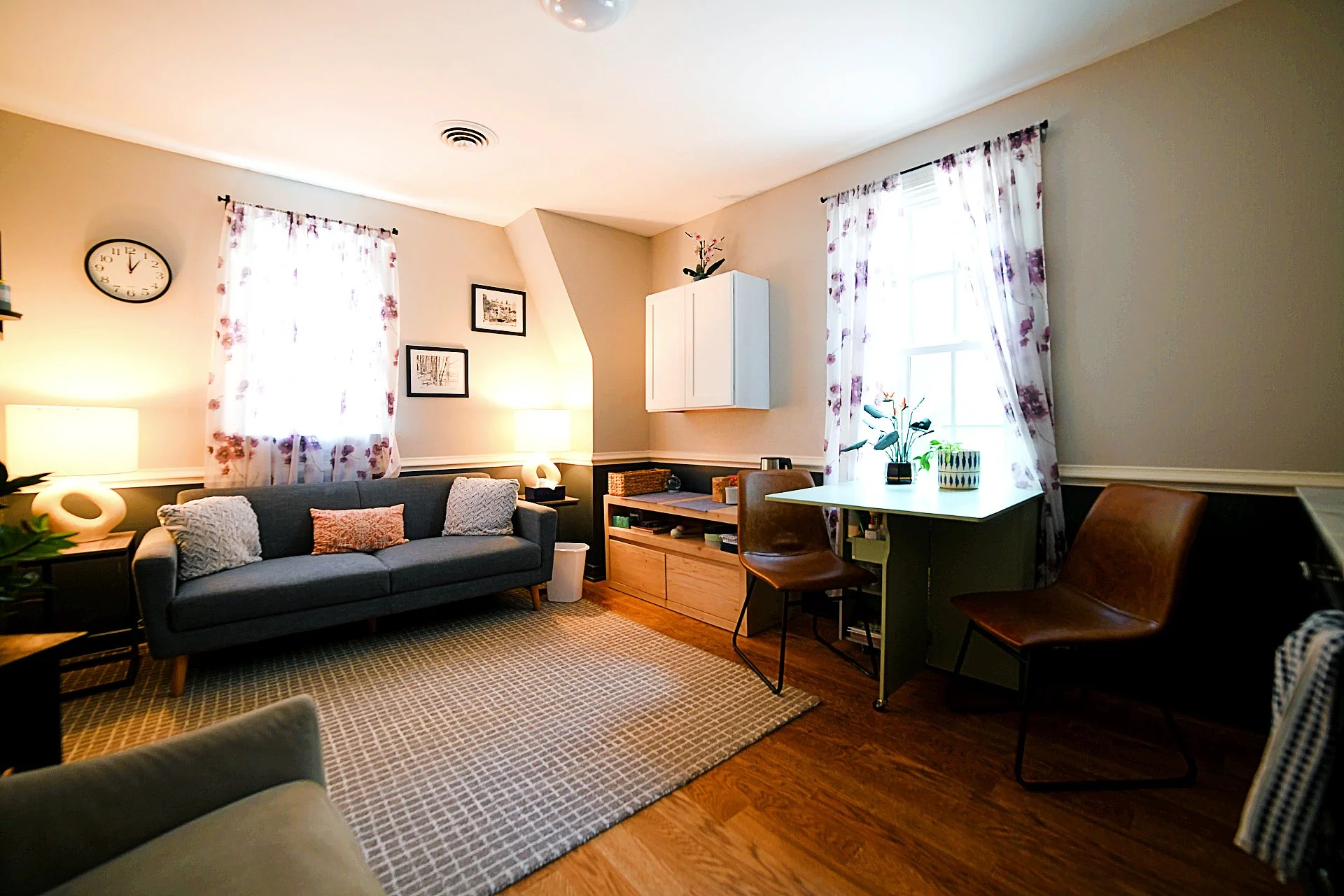 A living room with a gray sofa, decorative pillows, a side table with a lamp, two large windows with floral curtains, a white wall-mounted cabinet, a wooden TV stand, and a small desk with chairs and indoor plants.