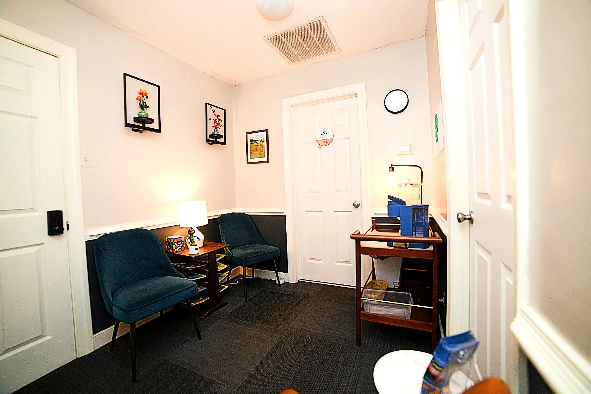 A small waiting room with three blue chairs, a small wooden table with a lamp and decorative items, and framed artwork on the walls. There is a white door in the background and a small table with blue folders on it. The room has white walls and a dark carpeted floor.