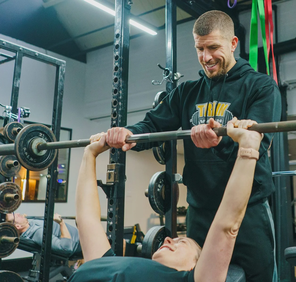 A man assisting a woman with a bench press in a gym, with weights and exercise equipment around.
