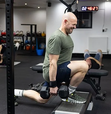 A man performing a fitness exercise in a gym, smiling while holding dumbbells, with a woman lying on a workout bench in the background.