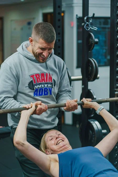 A woman performing a bench press exercise with a barbell at the gym, while a man spots her, both smiling.