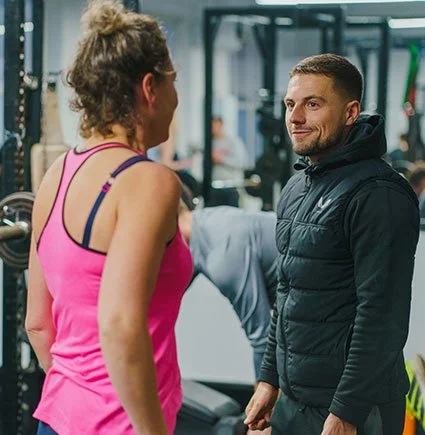 Two people having a conversation at a gym, with workout equipment in the background.