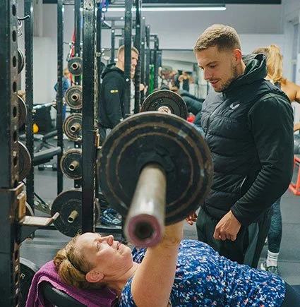 A woman lifting weights while lying on a bench in a gym, with a man spotting her, surrounded by gym equipment and other people working out.