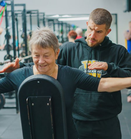 A young male trainer assisting an elderly woman during a workout at the gym.