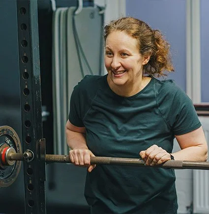A woman smiling while holding a barbell in a gym setting.