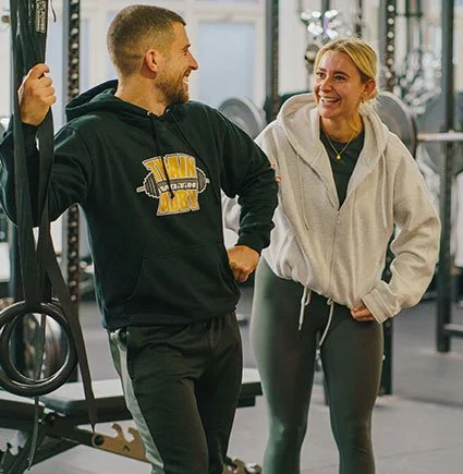 A man and a woman smiling and talking in a gym, dressed in athletic workout clothes.