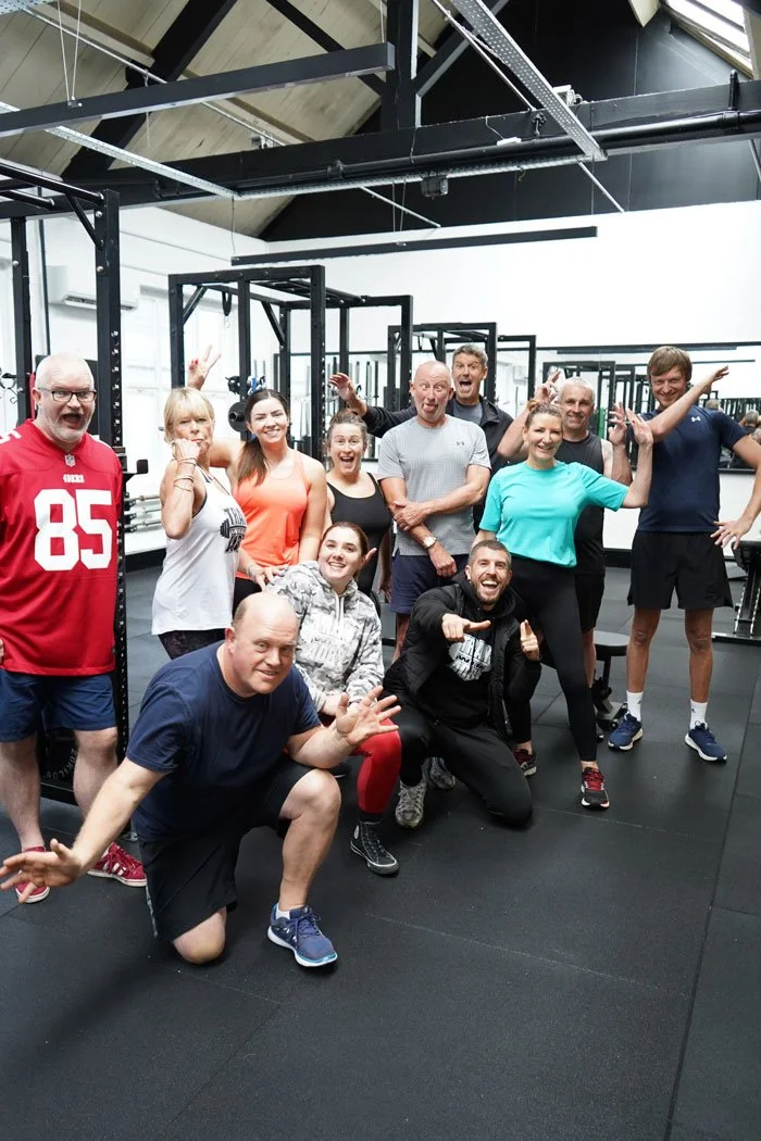 A group of ten people smiling and posing in a gym, some giving thumbs up or making playful gestures, with workout equipment in the background.