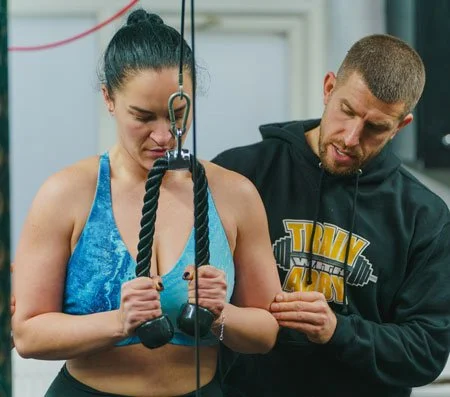 A woman using a cable machine for strength training, with a trainer guiding her and adjusting the settings.