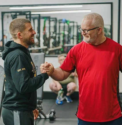 Two men shaking hands and smiling in a gym with workout equipment in the background.