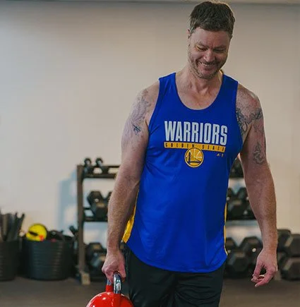 Man wearing a blue Golden State Warriors tank top, smiling in a gym.