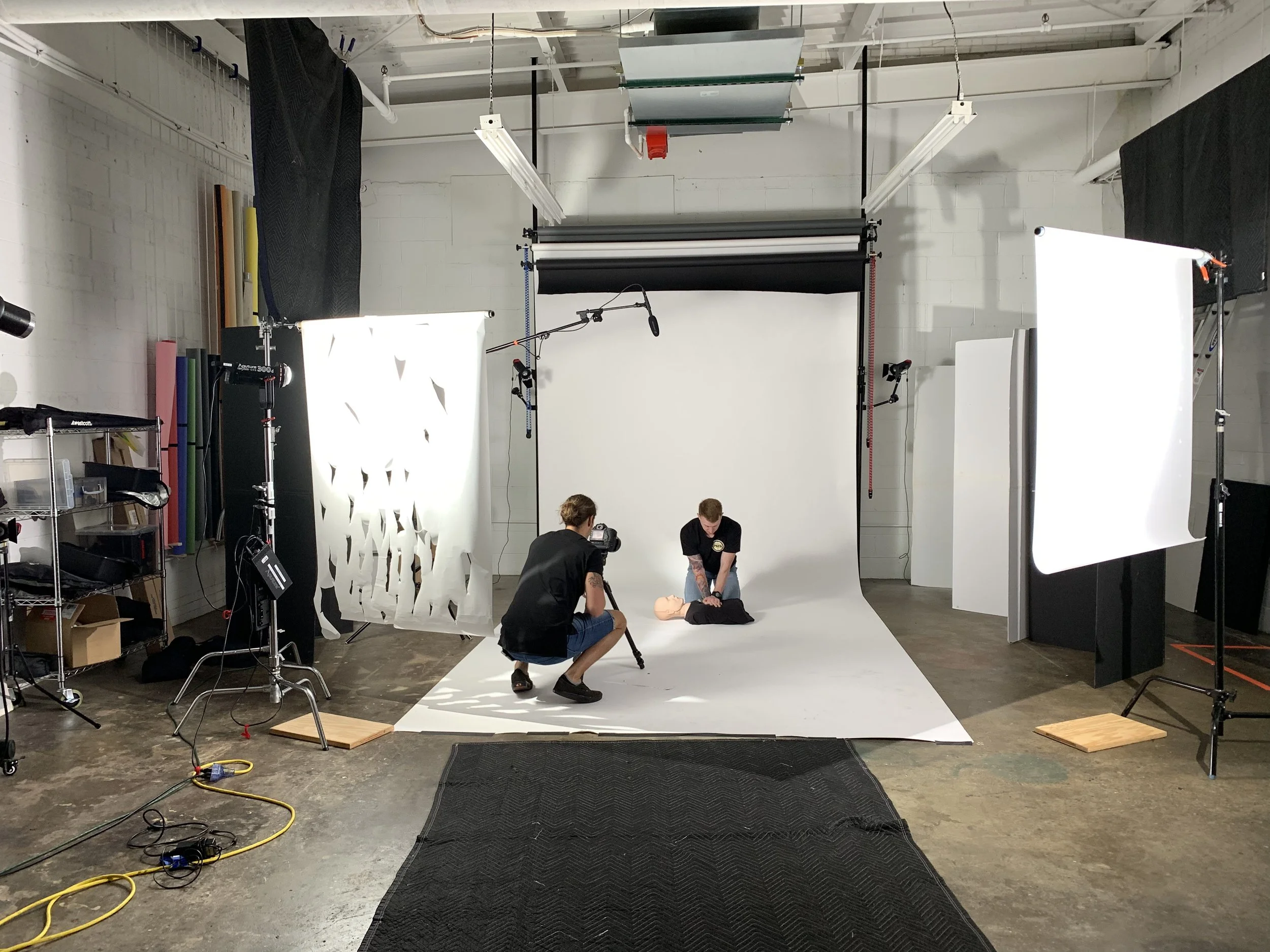 Photography studio setup with white seamless background, studio lighting, and a pair of people preparing to photograph a mannequin torso. Equipment includes softboxes, stands, and various modeling accessories, with studio crew working on the scene.