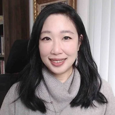 A woman with black hair wearing a gray turtleneck sweater smiling at the camera indoors.