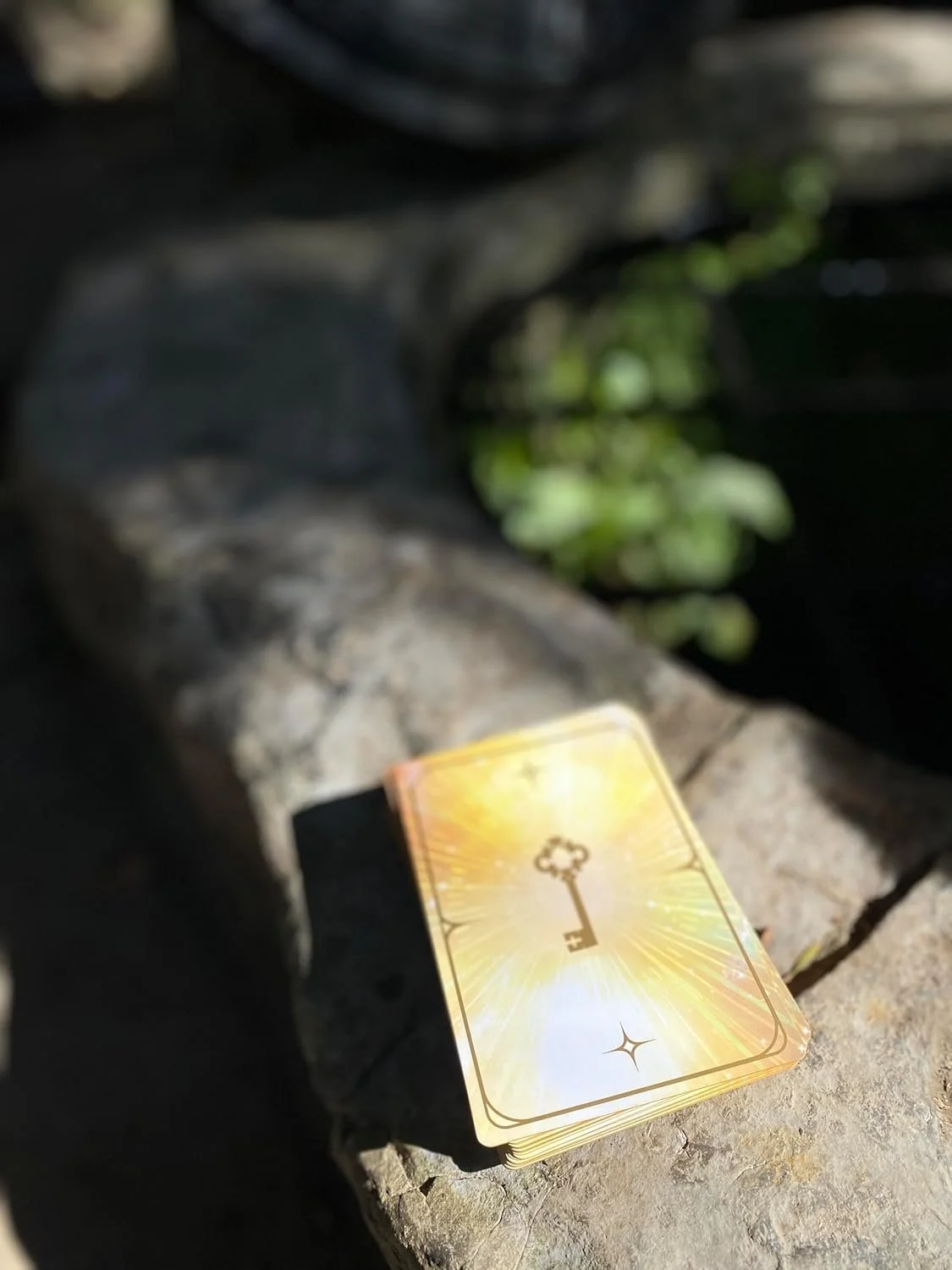 A deck of tarot or oracle cards with a golden key on the top card, placed on a stone surface outdoors with greenery in the background.