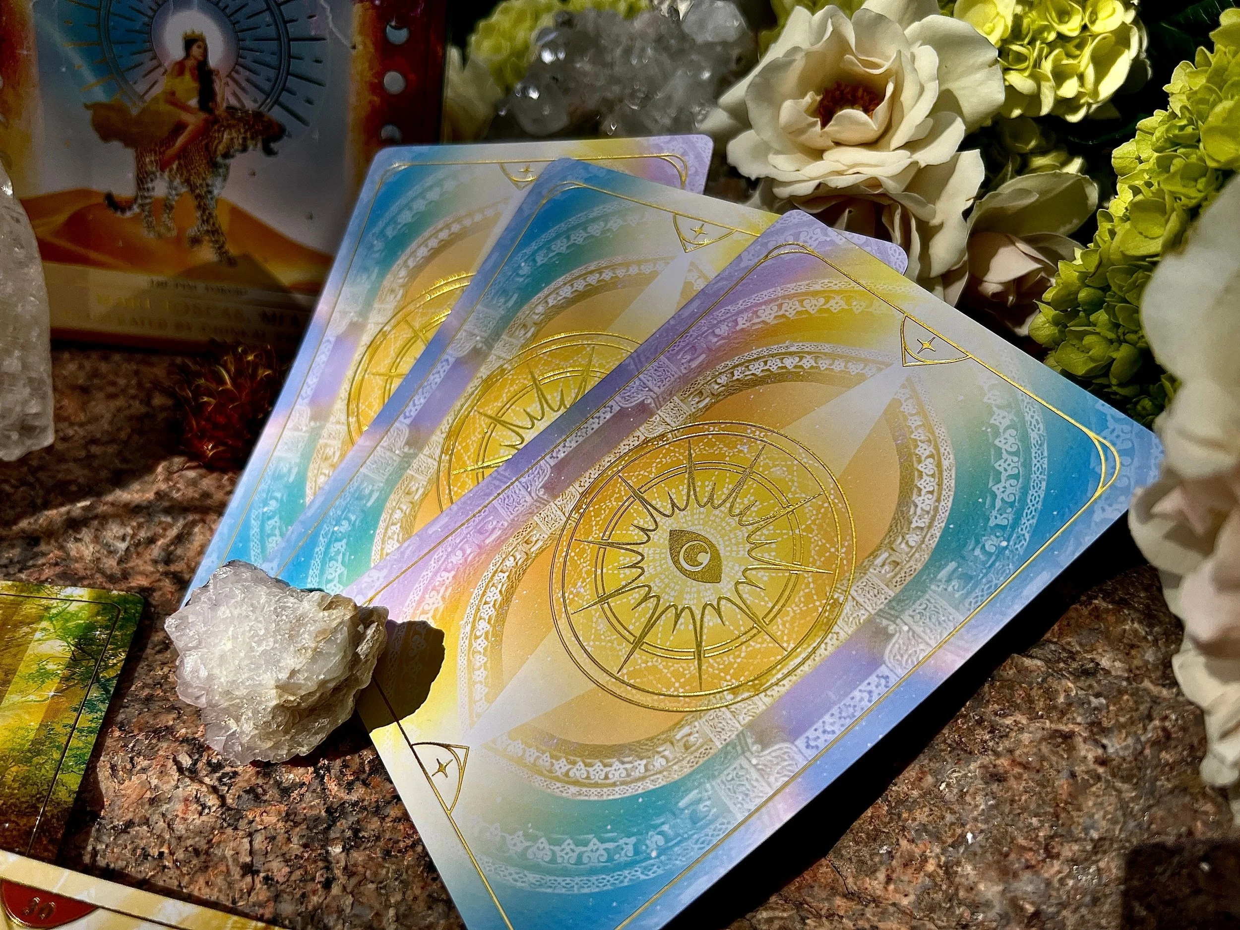 Three tarot cards with golden and pastel colors on a marble surface, surrounded by white and green flowers, a crystal, and a box with a figure riding a leopard on the background.