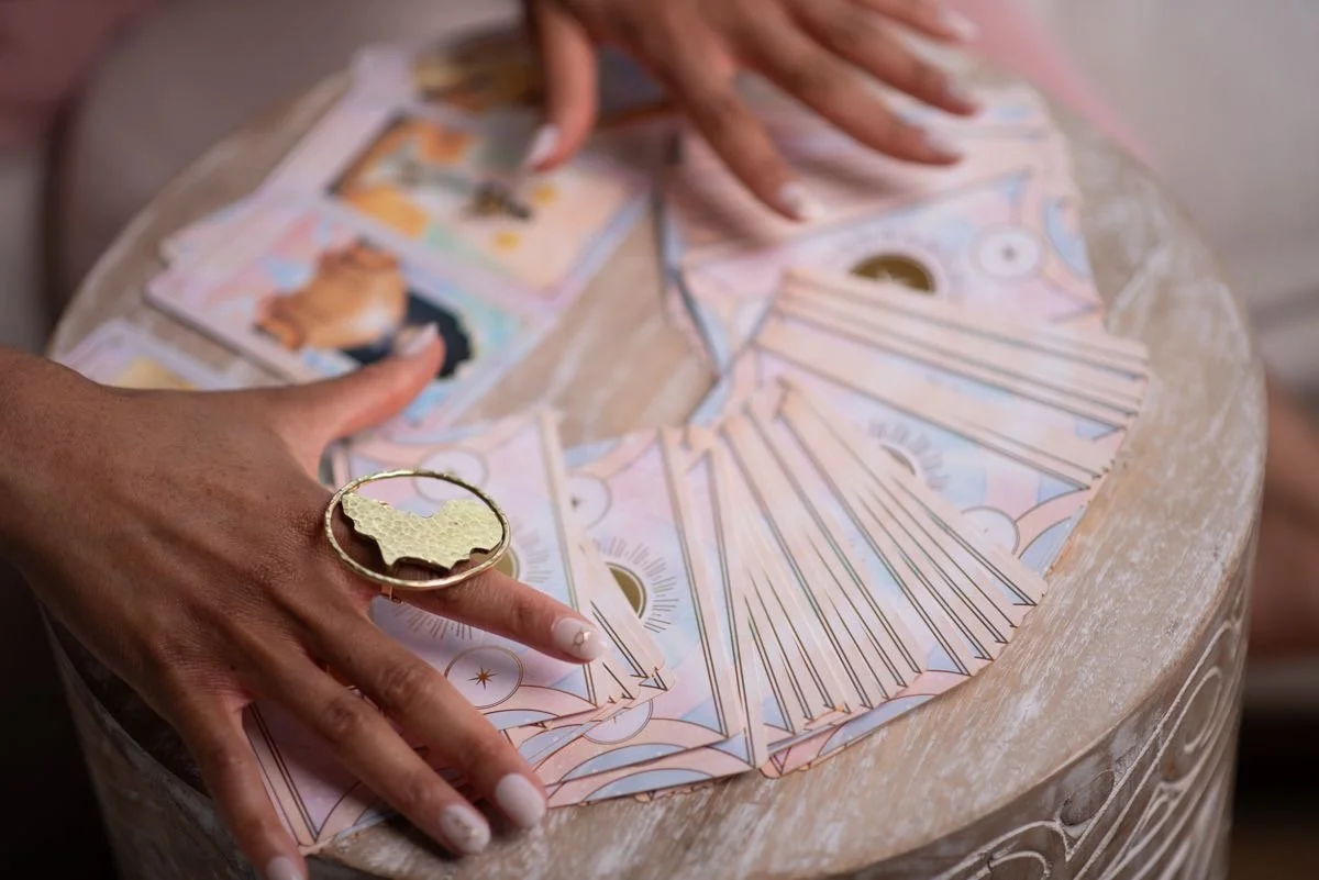 A person's hands are spread out on a round table covered with tarot cards, some of which have colorful illustrations. The person is wearing a large gold ring with an outline of the United States on their finger, and their nails are painted white with