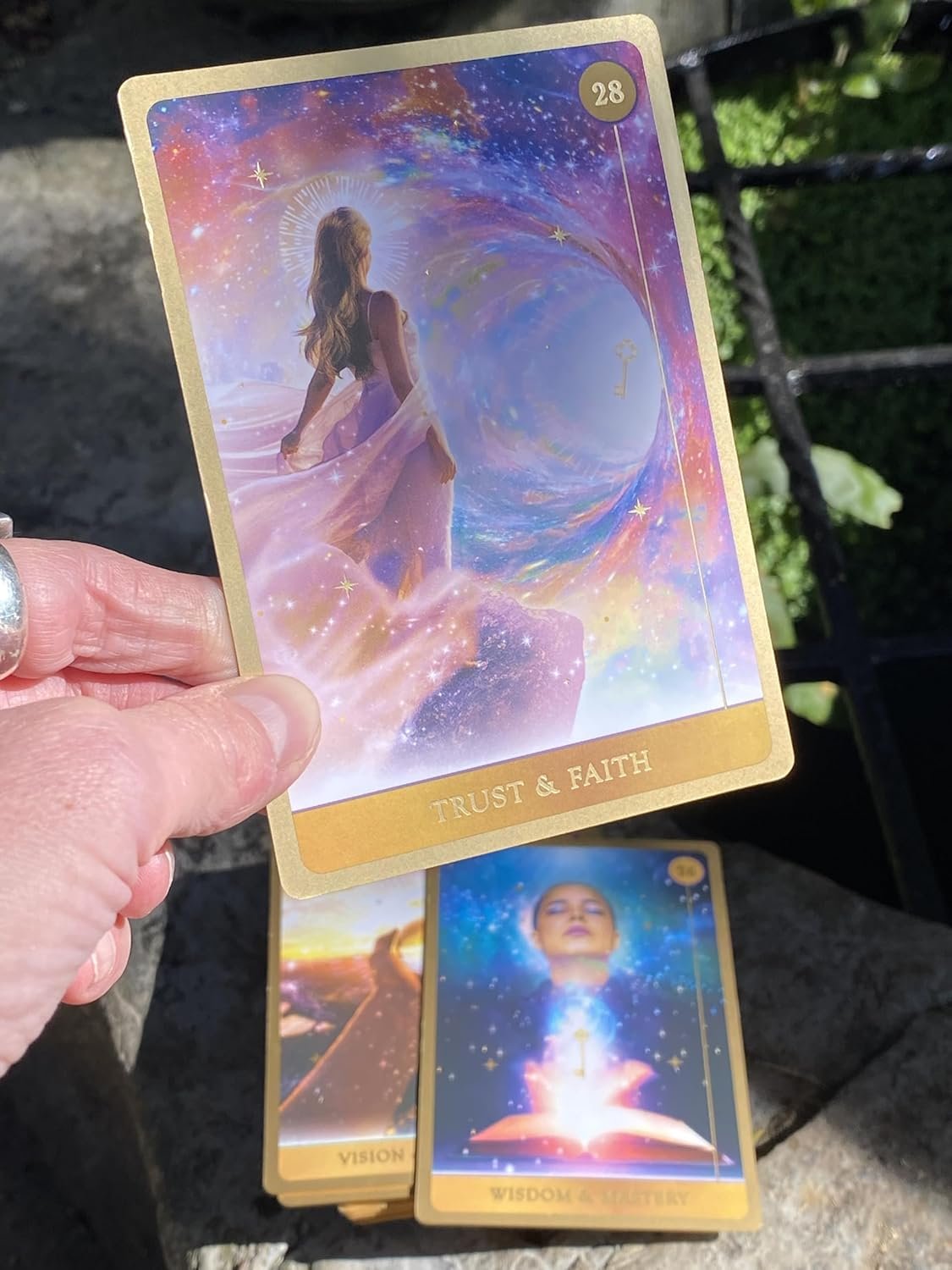 A person holding a card titled "TRUST & FAITH" with a woman in a flowing dress looking into a colorful, cosmic vortex with stars and a key. There are additional tarot cards on a fountain or stone surface outdoors.
