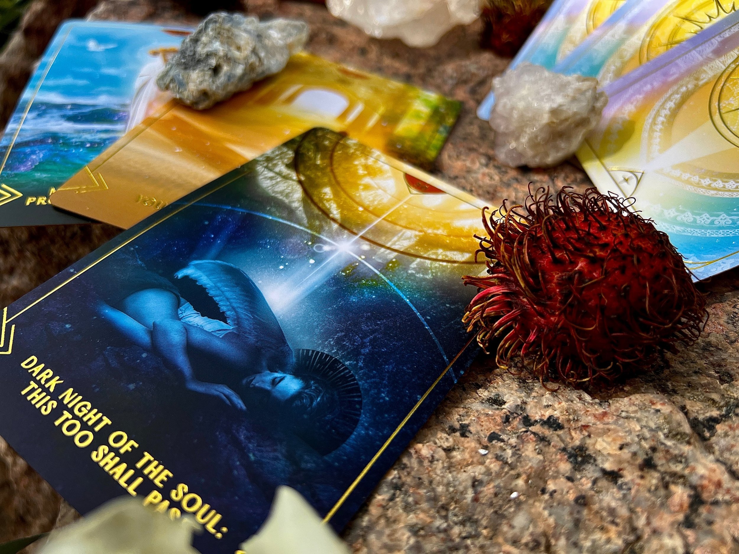 A collection of tarot cards, a red rambutan fruit, and some crystals arranged on a granite surface.