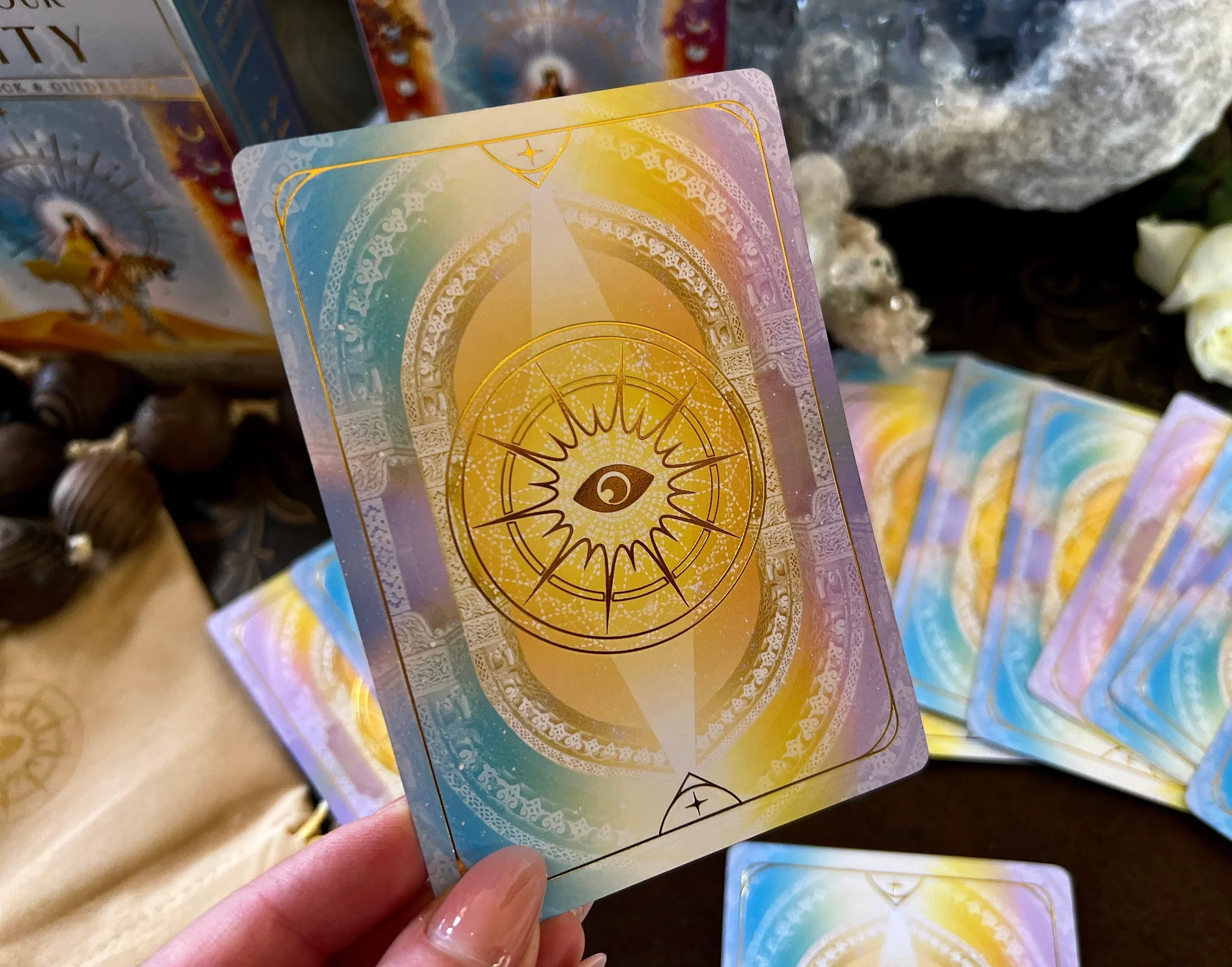A tarot card with a golden eye surrounded by rays and decorative patterns, featuring a color gradient of pastel yellow, purple, pink, and blue.