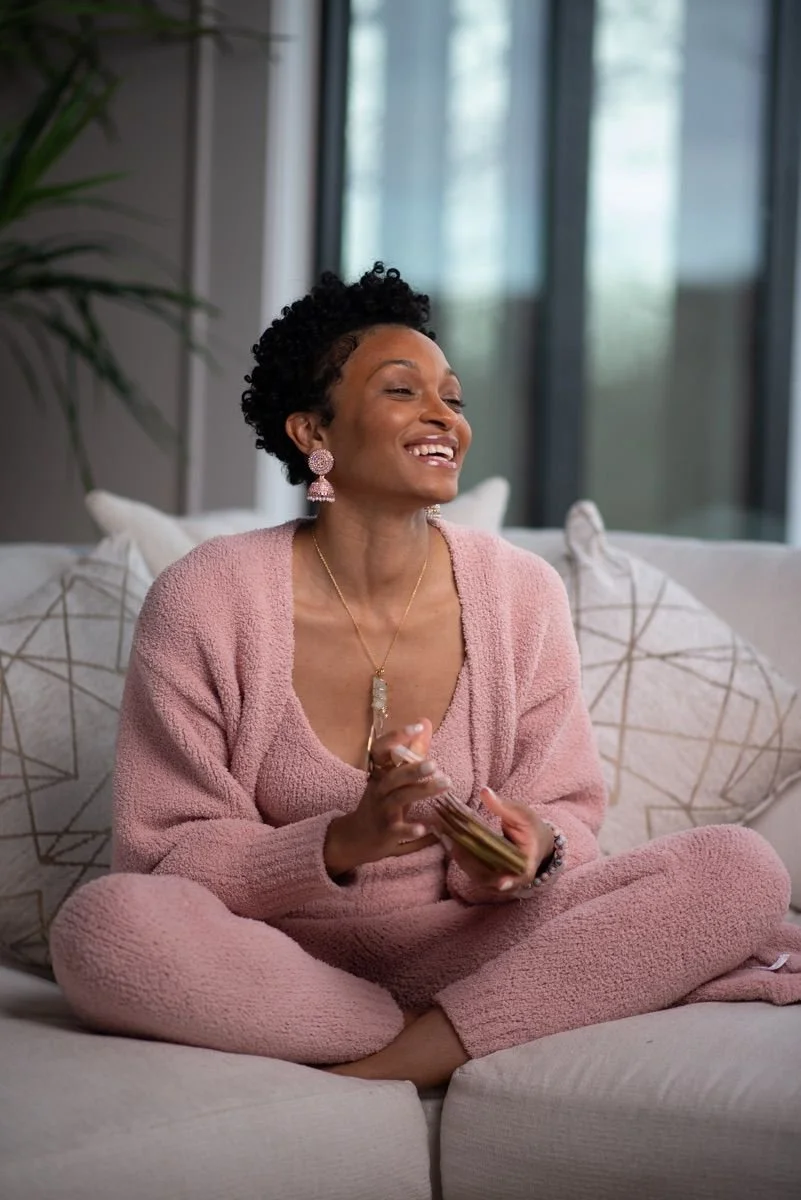 A woman sitting cross-legged on a sofa, smiling and holding a smartphone, inside a bright room with large windows and decorative pillows.
