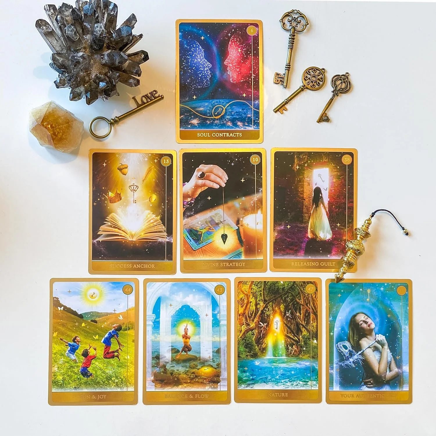 A collection of tarot or oracle cards laid out on a white surface, accompanied by decorative keys, a crystal cluster, and a gold-colored ornament. The cards feature mystical and nature-themed imagery and titles like "Soul Contracts," "Success Anchor,