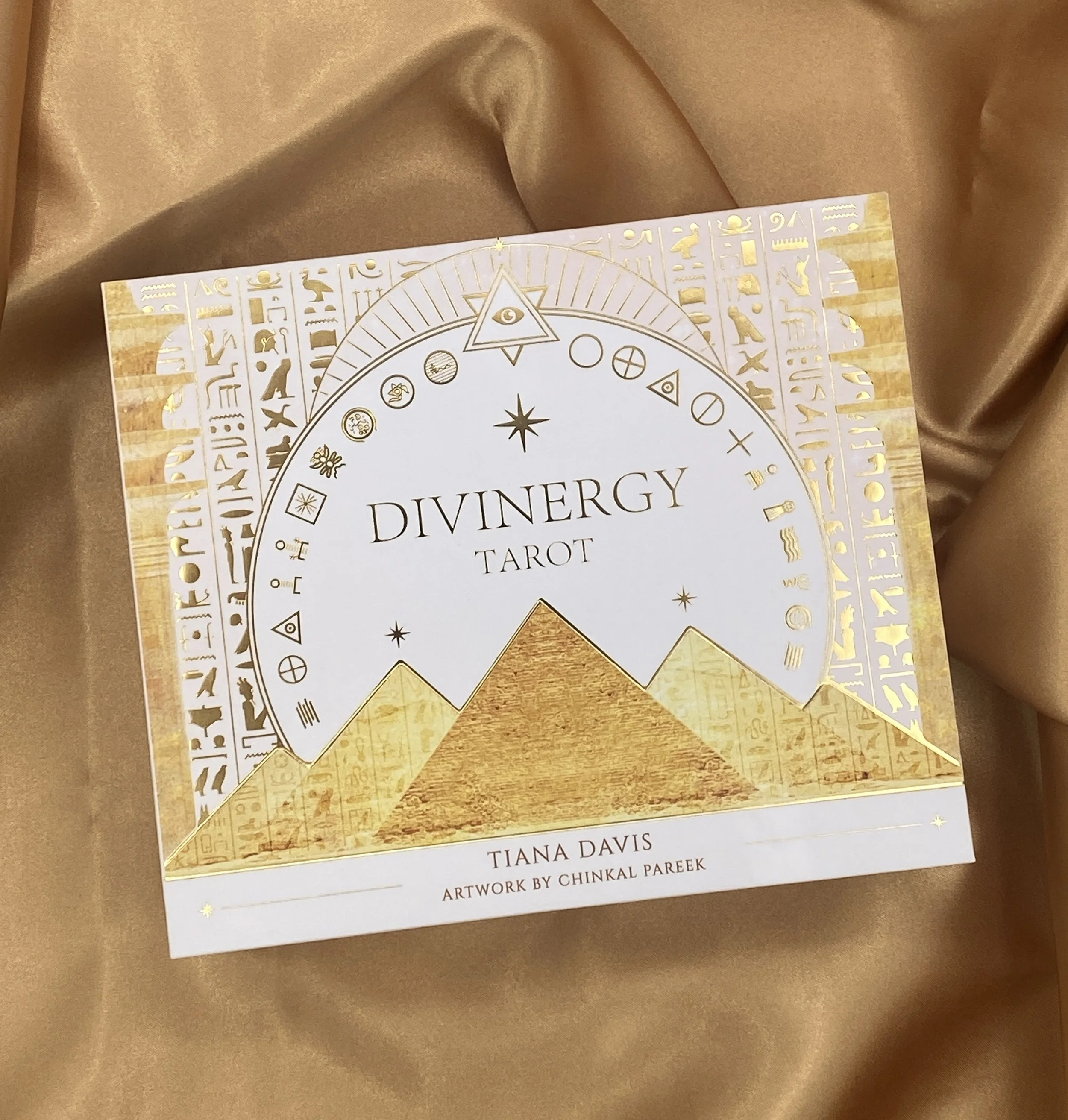 A tarot card deck box titled 'Divinergy Tarot' with gold and white design, featuring pyramids and symbols, against a gold satin background.
