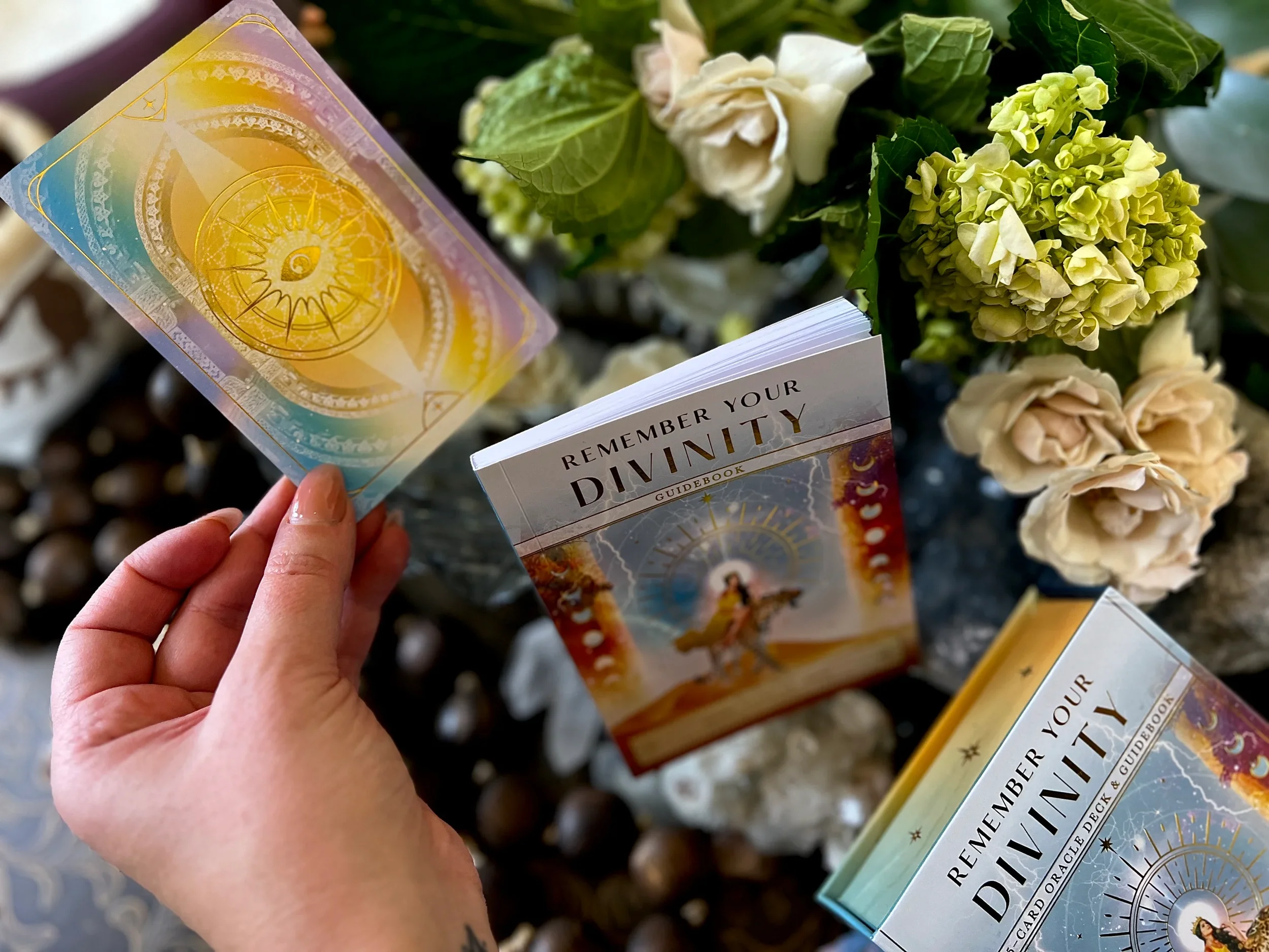A hand holding a deck of tarot cards with a colorful design, surrounded by flowers and open guidebook titled 'Remember Your Divinity' amid outdoor plants and stones.