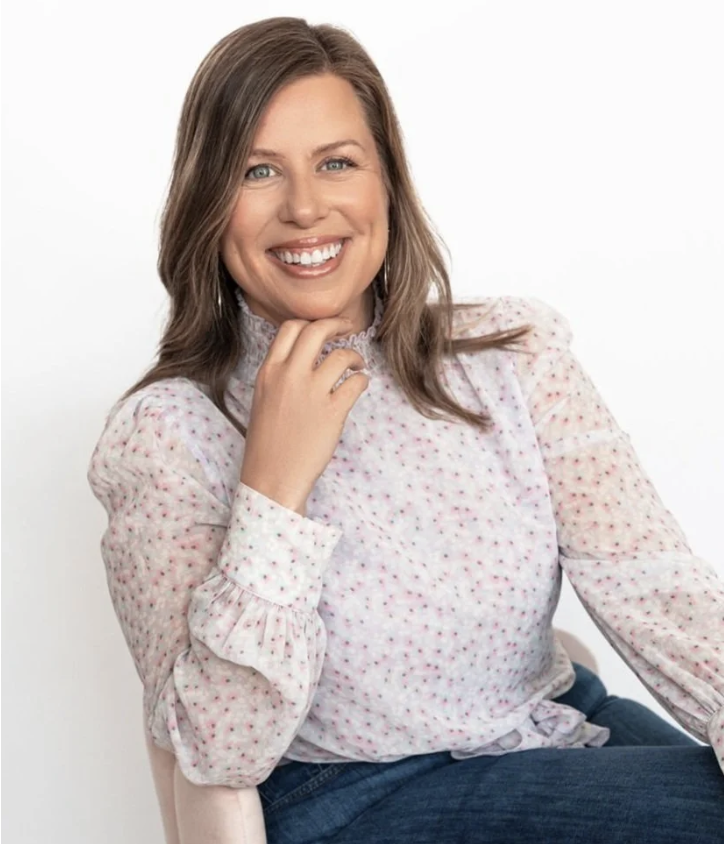 A woman with shoulder-length brown hair, smiling, wearing a white blouse with pink polka dots and blue jeans, sitting against a plain white background.