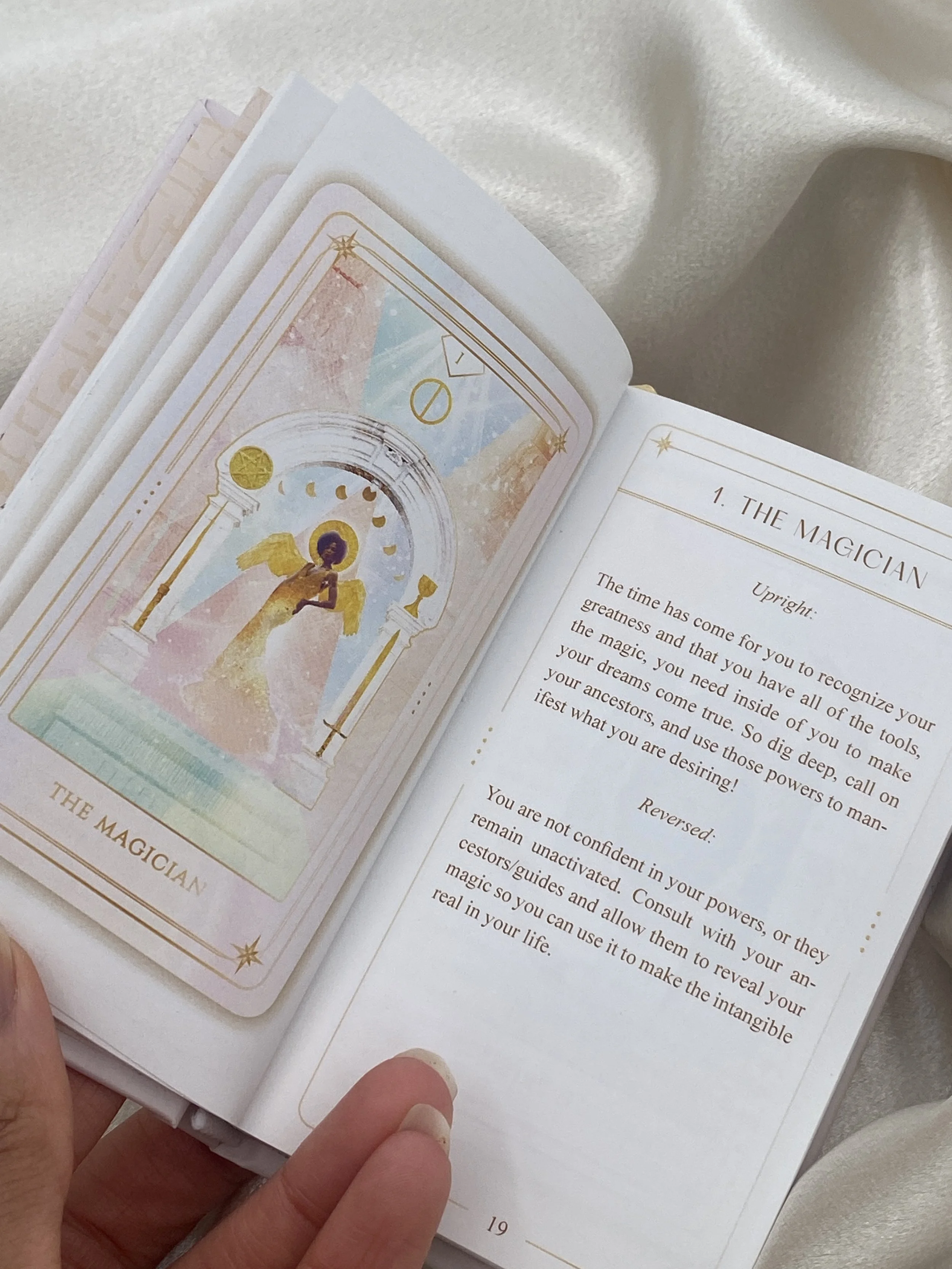 Open book showing a card and a page with text about The Magician, featuring a colorful illustration of a figure with angel wings behind a decorative arch.