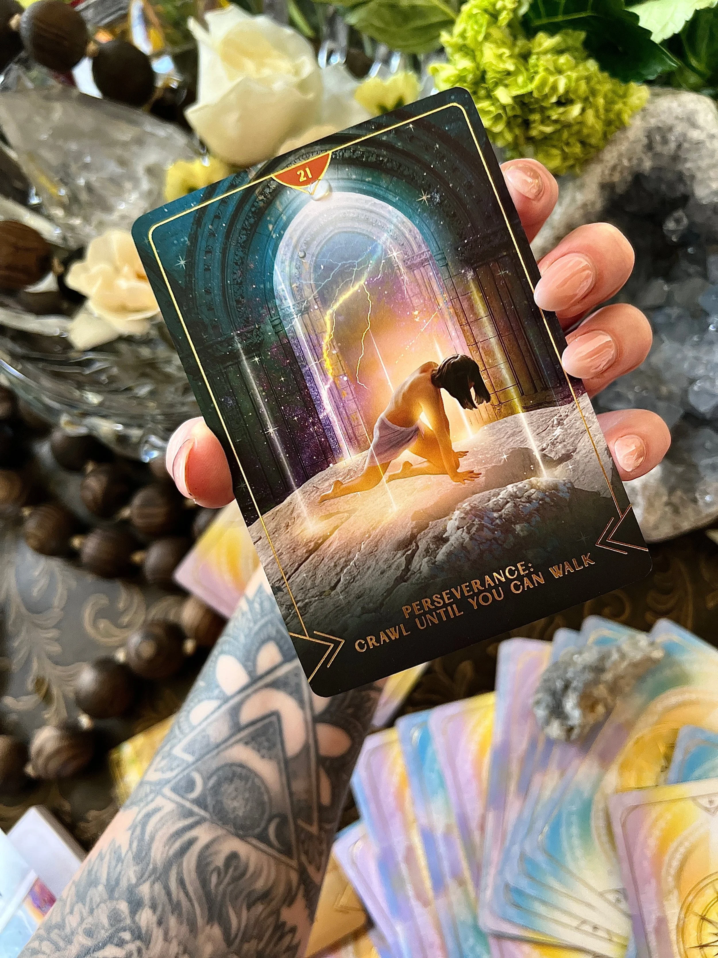 A person with a tattooed arm holding a tarot card featuring a girl in a yoga pose, with a mystical background of an arched door, lightning, and bright light. The card has the words 'Perseverance: Crawl Until You Can Walk' at the bottom.