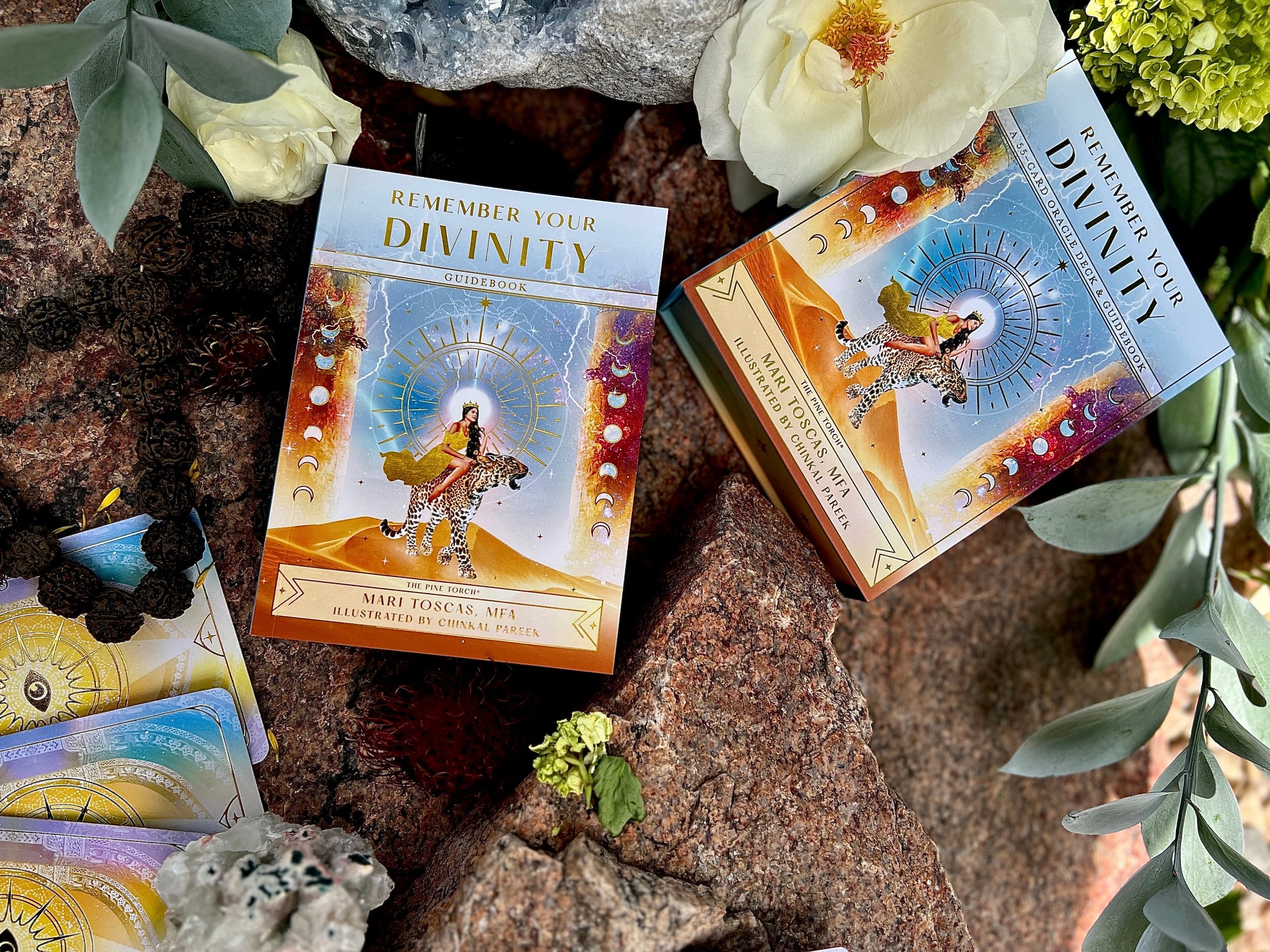 Deck of oracle cards titled "Remember Your Divinity" displayed on a rock with plants around, featuring an illustration of a woman riding a cheetah with a halo of celestial symbols.