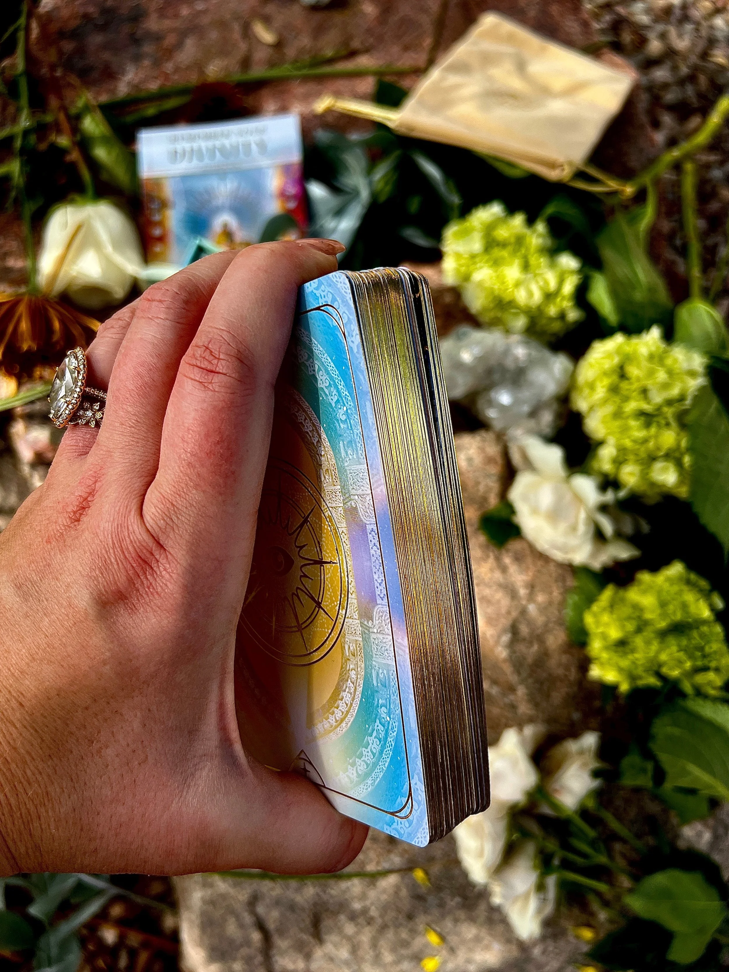 A hand holding a deck of tarot cards over a floral and rock background with flowers, crystals, and a white rose.