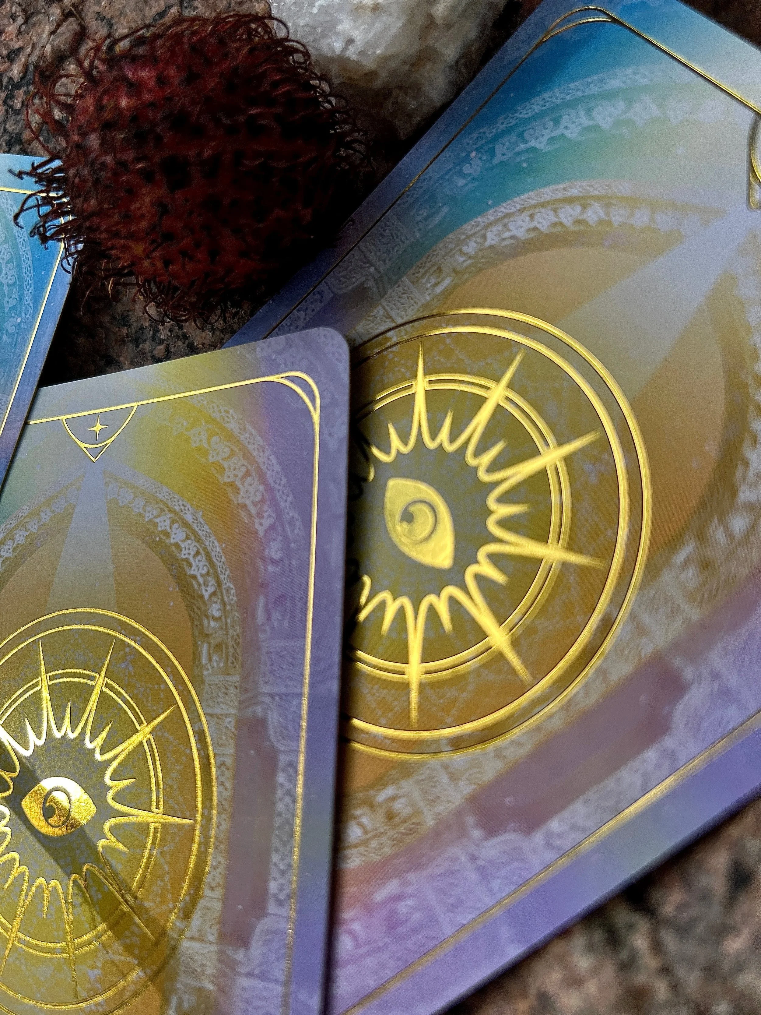 Close-up of two decks of tarot or oracle cards with a spiral and eye design in gold on the back, placed on a textured surface with a red, textured scrubby pad and a box with pastel-colored design in the background.