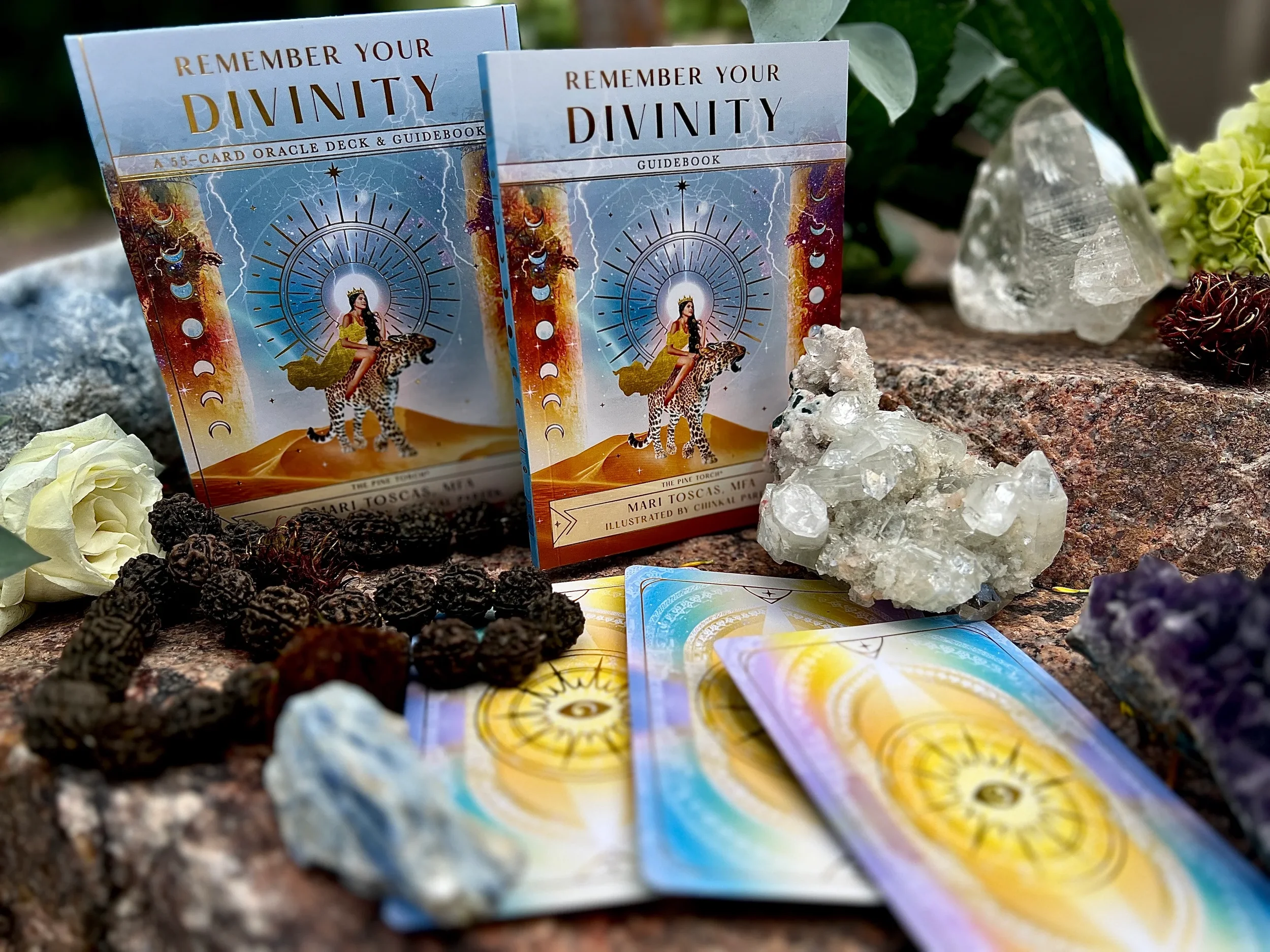 A collection of tarot and oracle cards titled "Remember Your Divinity" along with crystals, lava beads, and flowers on a rocky surface, arranged for a spiritual or tarot reading setup.