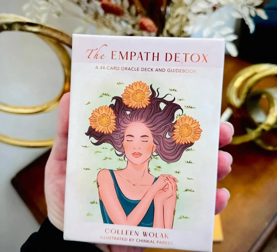 Cover of The Empath Detox, a card deck and guidebook, showing an illustration of a woman with closed eyes, long flowing hair decorated with orange flowers, wearing a blue top, holding her own hands. Background includes some small grass-like details.