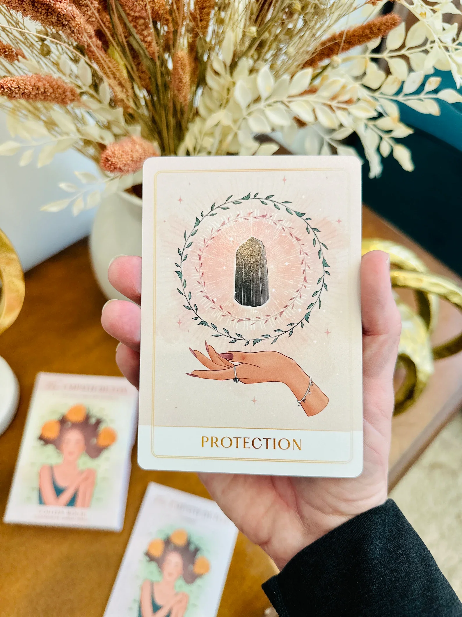 Tarot card titled 'Protection' featuring an illustrated hand with jewelry holding a black crystal surrounded by a circular wreath, with dried flowers in the background.