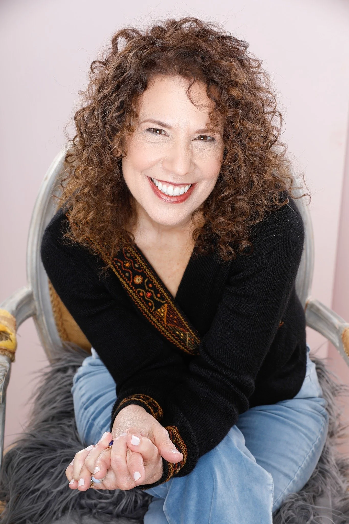 A woman with curly red hair smiling, sitting on a furry stool, wearing a black sweater with embroidered patterns and sitting cross-legged with her hands clasped.