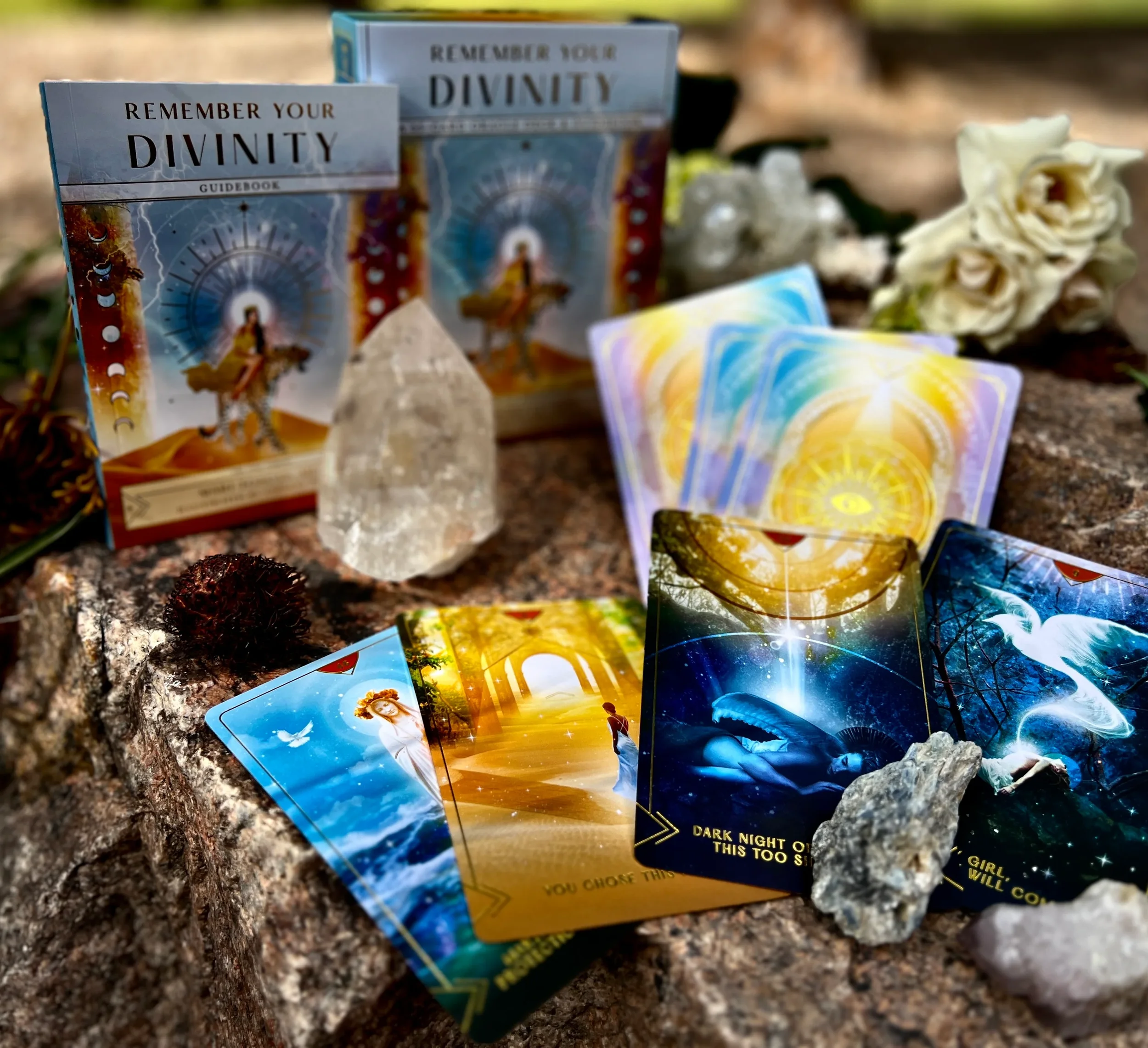 A collection of spiritual and tarot cards, a clear quartz crystal, white roses, a pinecone, and other natural items arranged on a rock surface.