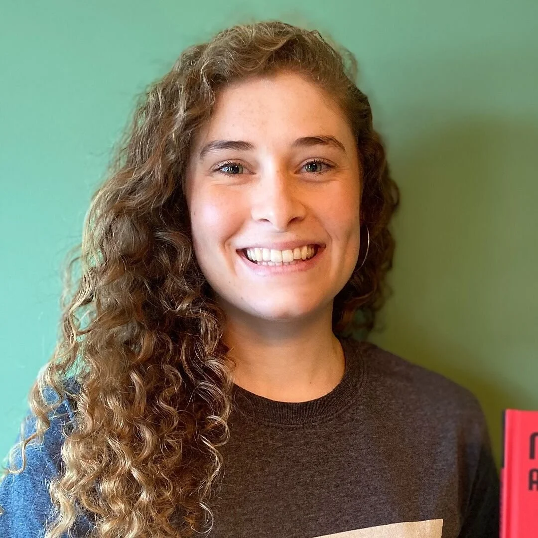 A young woman with curly hair, smiling, wearing a dark shirt, standing against a green background.