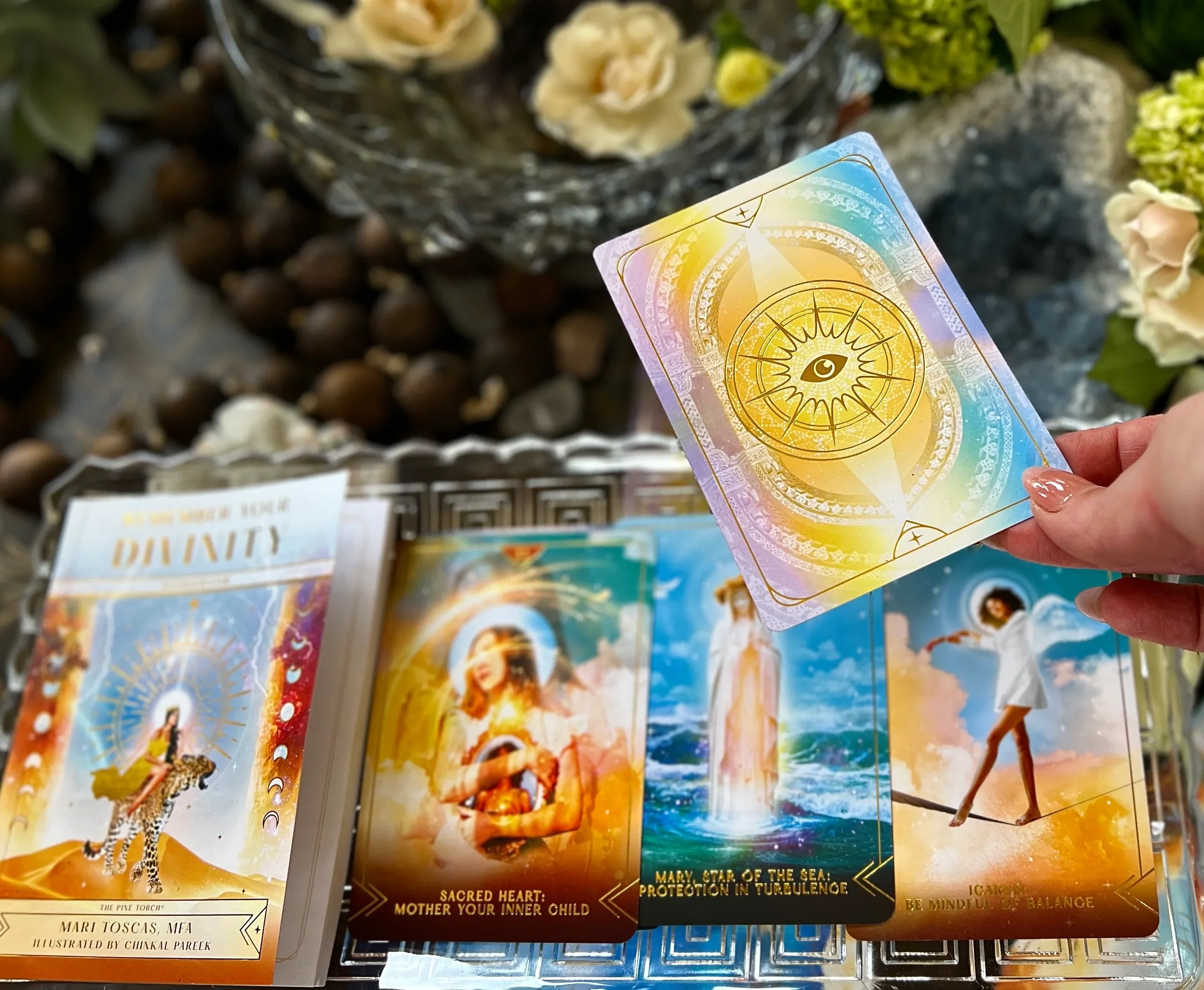 A person holds a card with an eye and celestial symbols, with other tarot cards spread out on a table nearby, surrounded by flowers and decorative objects.