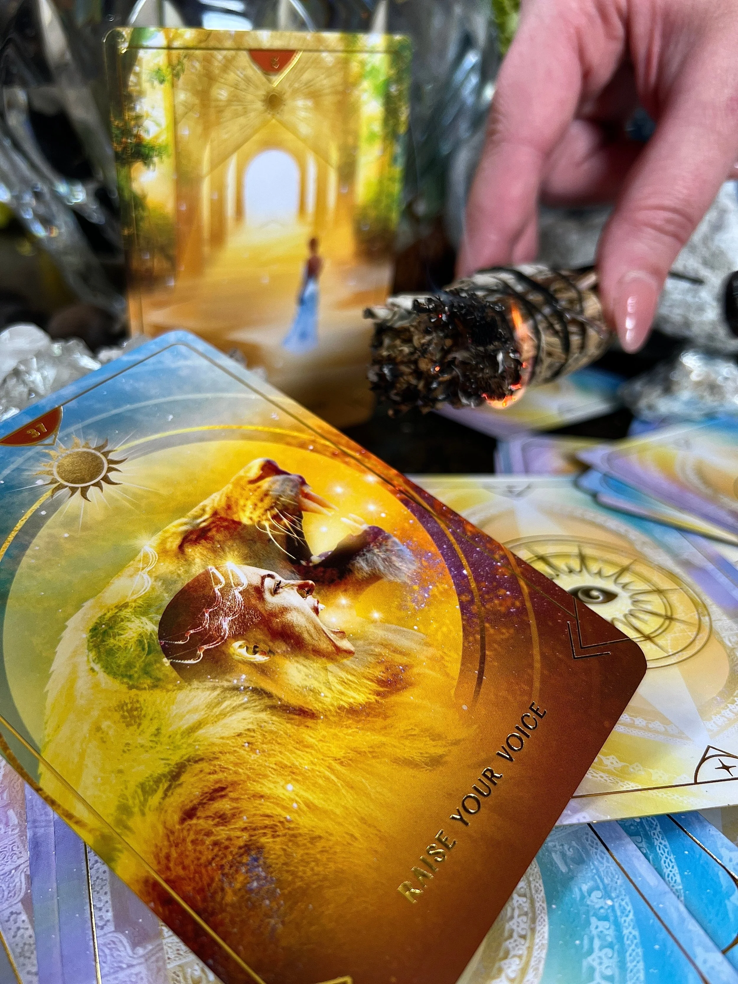 A person holding a burning sage stick above a table with colorful oracle cards, featuring spiritual and mystical images, including a woman, a lion, and a woman in blue, with the phrase "Raise Your Voice" on one card.