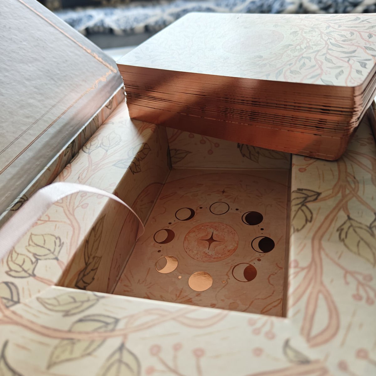 A box of tarot or oracle cards with moon phase illustrations and decorative floral pattern on the box interior.